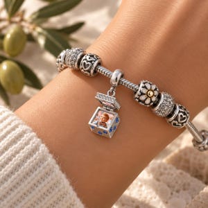 Puede incluir: Una pulsera de dijes de plata con un dije central en forma de relicario que se abre para mostrar una peque&ntilde;a fotograf&iacute;a de un ni&ntilde;o. El relicario est&aacute; decorado con piedras azules. La pulsera incluye otros dijes con corazones, cristales y una flor blanca.