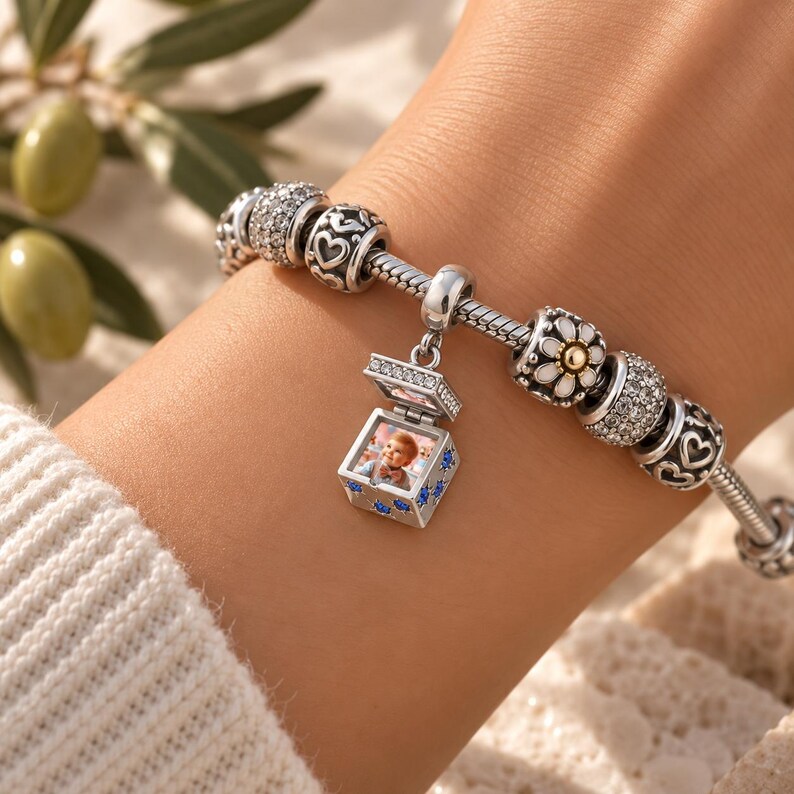 Puede incluir: Una pulsera de dijes de plata con un dije central en forma de relicario que se abre para mostrar una peque&ntilde;a fotograf&iacute;a de un ni&ntilde;o. El relicario est&aacute; decorado con piedras azules. La pulsera incluye otros dijes con corazones, cristales y una flor blanca.