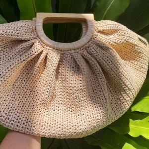 May include: A beige crocheted handbag with a wooden handle. The bag has a rounded shape with pleats at the top and a circular wooden handle. The bag is set against a background of green leaves.