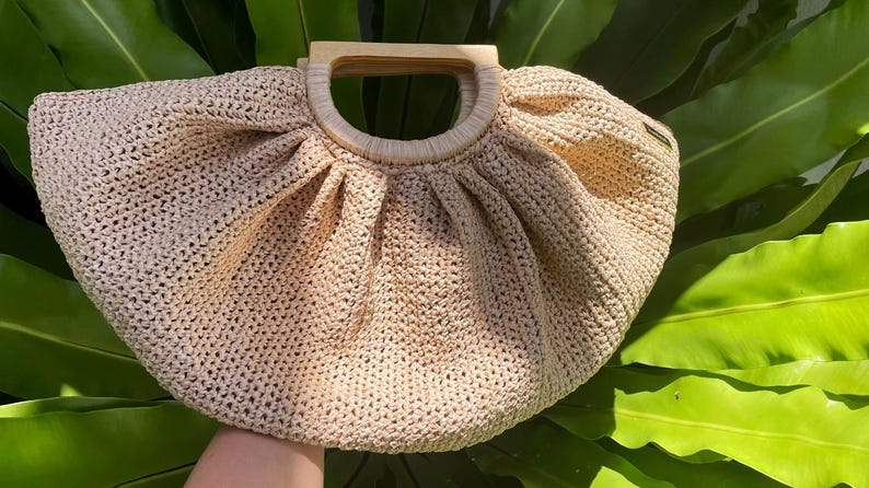 May include: A beige crocheted handbag with a wooden handle. The bag has a rounded shape with pleats at the top and a circular wooden handle. The bag is set against a background of green leaves.