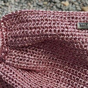 K&ouml;nnte beinhalten: Eine handgefertigte, staubrosa Handtasche mit gewebter Textur. Die Tasche hat einen Holzgriff und einen kreisf&ouml;rmigen, satinartigen Ring. Ein kleines Metallschildchen tr&auml;gt die Aufschrift "handmade".