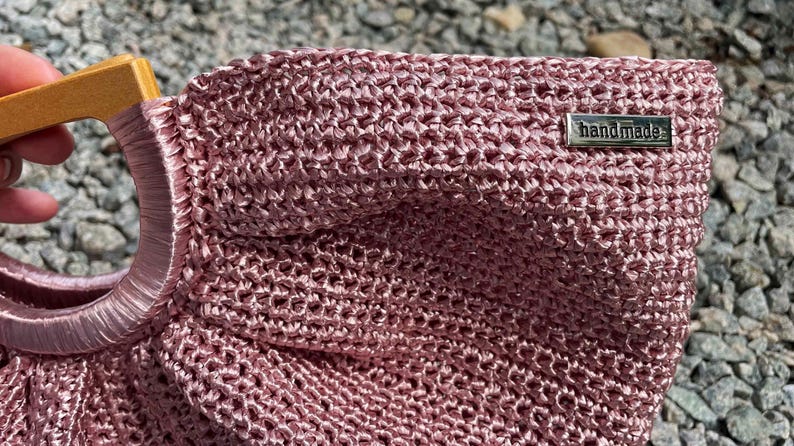 K&ouml;nnte beinhalten: Eine handgefertigte, staubrosa Handtasche mit gewebter Textur. Die Tasche hat einen Holzgriff und einen kreisf&ouml;rmigen, satinartigen Ring. Ein kleines Metallschildchen tr&auml;gt die Aufschrift "handmade".