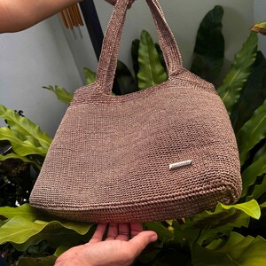 May include: A handmade, light brown tote bag with a woven texture. The bag has two handles and a small, rectangular metal tag that reads "handmade". The bag is held up against a background of green leaves.