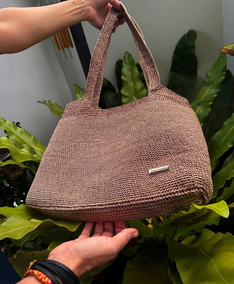 May include: A handmade, light brown tote bag with a woven texture. The bag has two handles and a small, rectangular metal tag that reads "handmade". The bag is held up against a background of green leaves.
