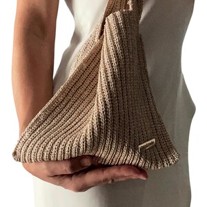 May include: A beige, handmade, knitted bag with a unique triangular shape. The bag has a ribbed texture and a small, rectangular tag that reads "handmade". The bag is held by a person wearing a white dress.