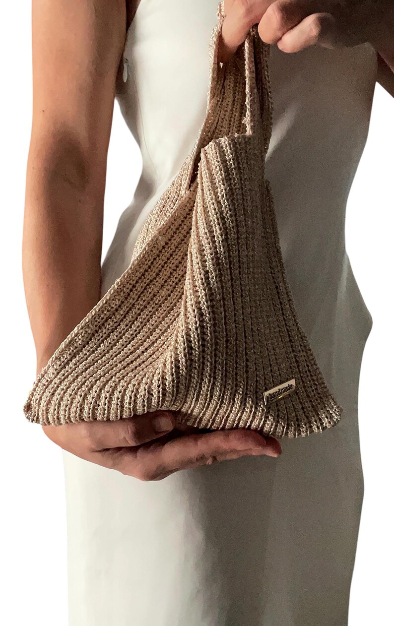 May include: A beige, handmade, knitted bag with a unique triangular shape. The bag has a ribbed texture and a small, rectangular tag that reads "handmade". The bag is held by a person wearing a white dress.