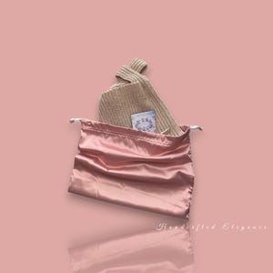 May include: A light pink satin drawstring bag with a beige knit hat inside. A small card with blue floral designs is visible. The background is a solid light pink color with the words "Handcrafted Elegance" in cursive.