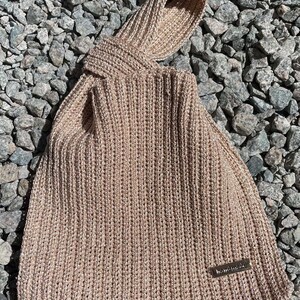 May include: A beige knitted tote bag with a knotted handle. The bag is made of a thick, textured yarn and features a small, rectangular "handmade" tag. The bag is resting on a bed of gray rocks.