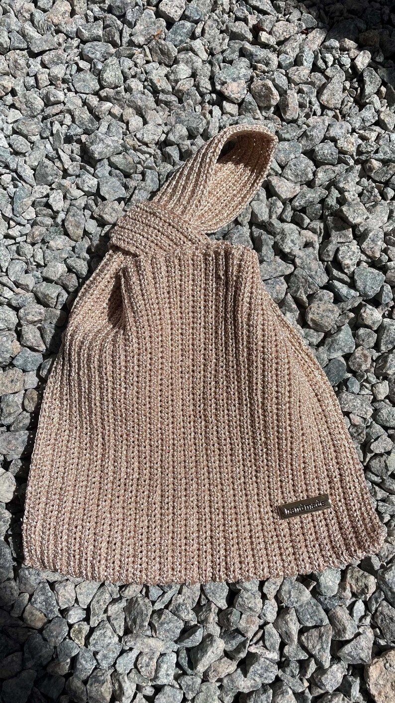 May include: A beige knitted tote bag with a knotted handle. The bag is made of a thick, textured yarn and features a small, rectangular "handmade" tag. The bag is resting on a bed of gray rocks.