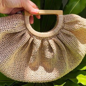 May include: A beige, crocheted handbag with a wooden handle. The bag has a rounded shape with a pleated design. The handle is a light brown color. The bag is set against a background of green leaves.