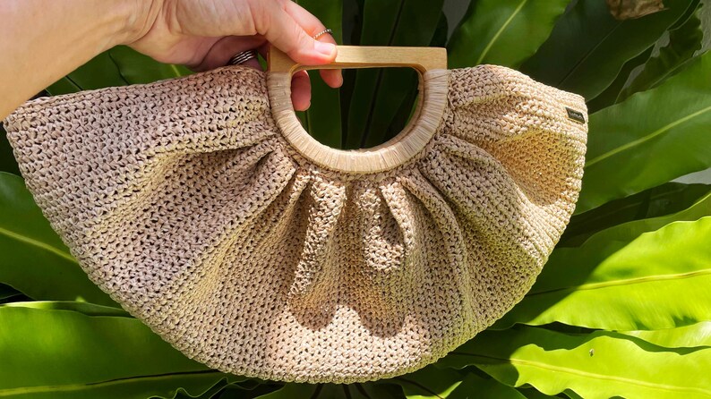 May include: A beige, crocheted handbag with a wooden handle. The bag has a rounded shape with a pleated design. The handle is a light brown color. The bag is set against a background of green leaves.
