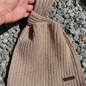 May include: A beige knitted bag with a knotted handle. The bag has a rectangular shape with a small rectangular tag that reads "handmade". The bag is made of a textured, ribbed knit material.