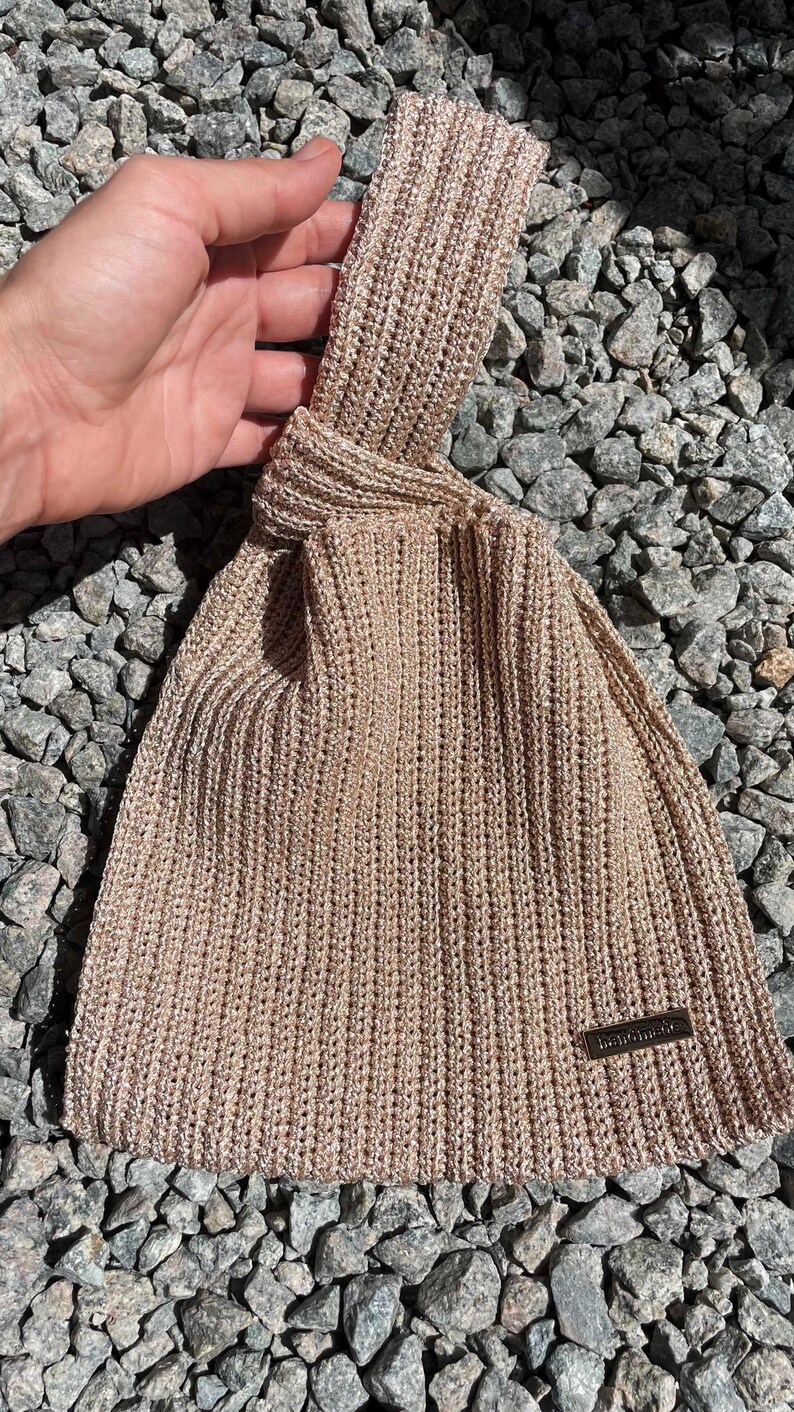 May include: A beige knitted bag with a knotted handle. The bag has a rectangular shape with a small rectangular tag that reads "handmade". The bag is made of a textured, ribbed knit material.