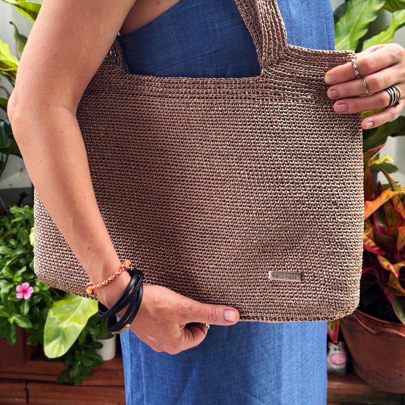 May include: A brown, handmade crochet tote bag with a rectangular shape and two handles. The bag has a small metal tag that reads "handmade". The bag is being held by a person wearing a blue dress.