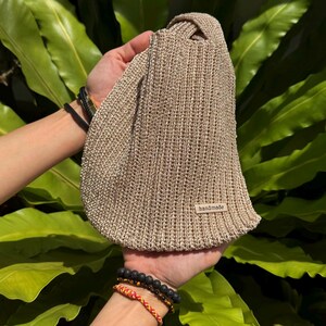 May include: A beige, handmade knitted bag with a knotted handle. The bag is held up by two hands. A small tag that says "handmade" is sewn on the bag. The background is a lush green plant.