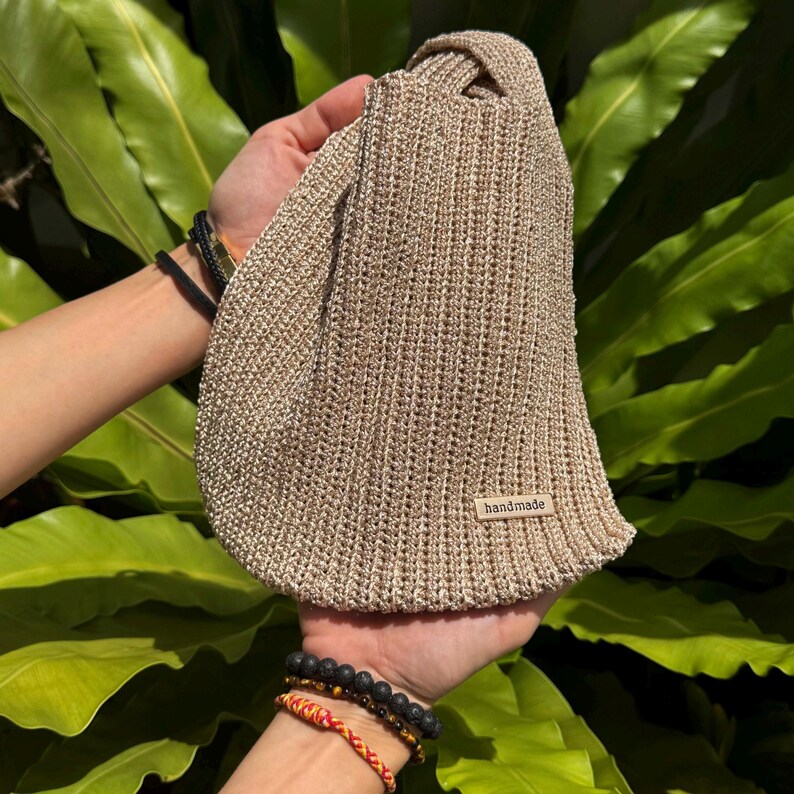 May include: A beige, handmade knitted bag with a knotted handle. The bag is held up by two hands. A small tag that says "handmade" is sewn on the bag. The background is a lush green plant.