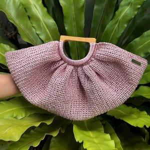 K&ouml;nnte beinhalten: Eine rosa gewebte Handtasche mit Holzgriff. Die Tasche hat eine abgerundete Form und eine strukturierte Oberfl&auml;che. Der Griff ist hellbraun und oben an der Tasche befestigt. Die Tasche ist vor einem Hintergrund aus gr&uuml;nen Bl&auml;ttern platziert.