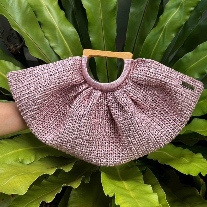 K&ouml;nnte beinhalten: Eine rosa gewebte Handtasche mit Holzgriff. Die Tasche hat eine abgerundete Form und eine strukturierte Oberfl&auml;che. Der Griff ist hellbraun und oben an der Tasche befestigt. Die Tasche ist vor einem Hintergrund aus gr&uuml;nen Bl&auml;ttern platziert.