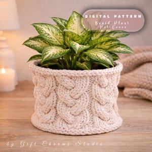 Puede incluir: Una funda de maceta de punto beige con un diseño de trenzas, que contiene una planta de hojas verdes y blancas. El texto "DIGITAL PATTERN Braid Plant Pot Cover" está en una pancarta marrón rosado. Las palabras "by Gift Charms Studio" están en la parte inferior.