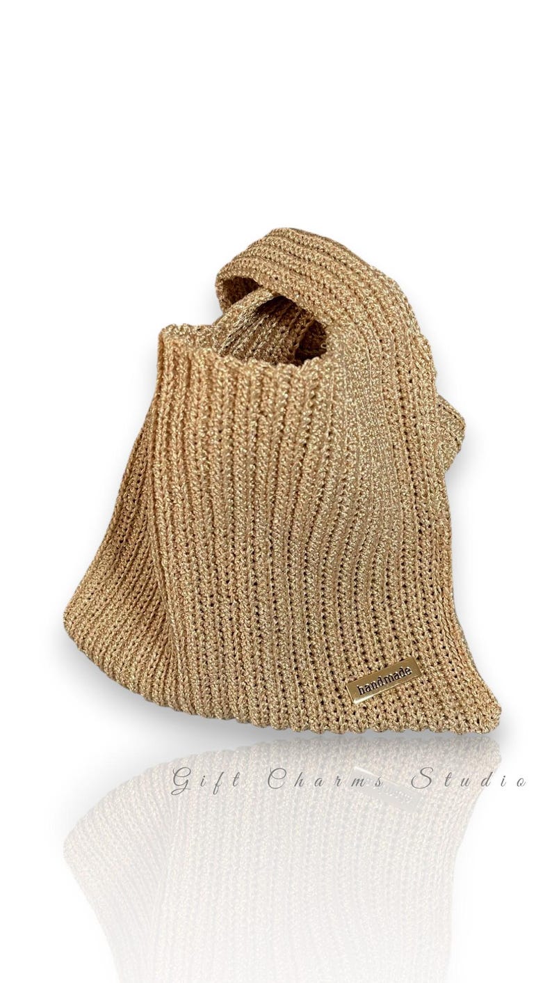 May include: A gold-colored, knitted tote bag with a handle. The bag has a rectangular shape with a wide opening and a small, rectangular tag that reads "handmade". The bag is made of a shiny, textured material.
