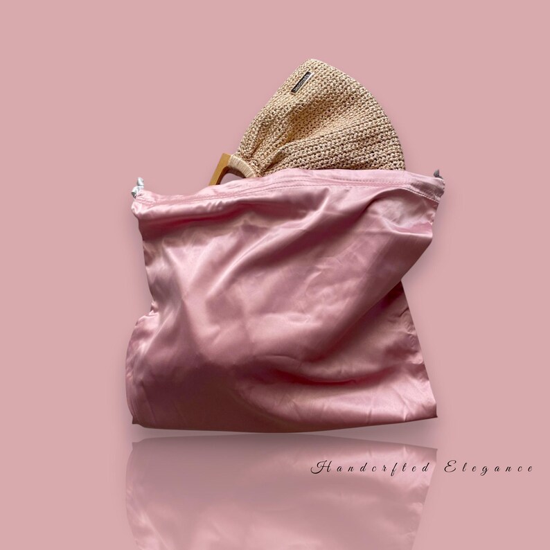 May include: A light pink satin bag with a beige woven handbag inside. The handbag has wooden handles. The background is a solid light pink color. The text "Handcrafted Elegance" is at the bottom.