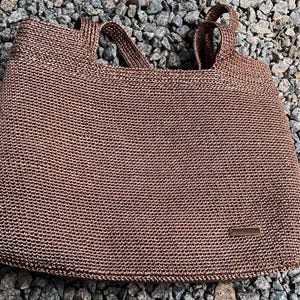 May include: A brown woven tote bag with two handles. The bag has a rectangular shape and a small rectangular tag on the front. The bag is sitting on a bed of small grey rocks.