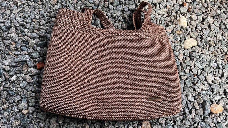 May include: A brown woven tote bag with two handles. The bag has a rectangular shape and a small rectangular tag on the front. The bag is sitting on a bed of small grey rocks.