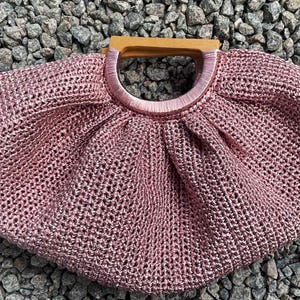 K&ouml;nnte beinhalten: Eine handgefertigte, altrosa geh&auml;kelte Handtasche mit Holzgriff. Die Tasche hat eine runde Form mit einem kreisf&ouml;rmigen Griff und einem kleinen Etikett an der Seite. Die Tasche besteht aus einem gewebten Material.