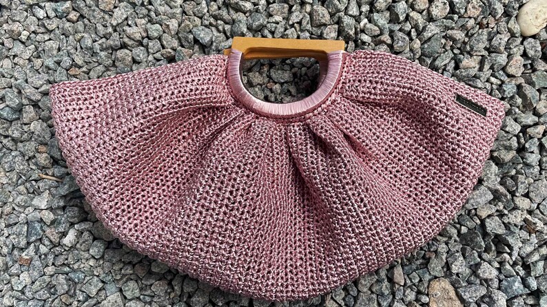 K&ouml;nnte beinhalten: Eine handgefertigte, altrosa geh&auml;kelte Handtasche mit Holzgriff. Die Tasche hat eine runde Form mit einem kreisf&ouml;rmigen Griff und einem kleinen Etikett an der Seite. Die Tasche besteht aus einem gewebten Material.