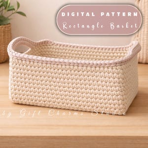 Op de afbeelding: Een rechthoekige, beige gehaakte mand met een gevlochten, lichtroze rand langs de bovenrand en handvatten. De tekst "DIGITAL PATTERN Rectangle Basket" wordt weergegeven in een roze ovaal. De woorden "by Gift Charms Studio" staan onderaan.