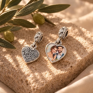 May include: Two heart-shaped charms with silver-toned metal settings. One charm features a swirling embossed pattern, while the other displays a colour photograph of a couple. Both charms have a bail accented with small, clear, sparkling stones.