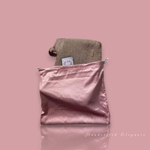 May include: A pink satin drawstring bag with a gold-coloured woven handbag. A small card is visible inside the handbag. The background is a solid pink colour. The words "Handcrafted Elegance" are at the bottom.