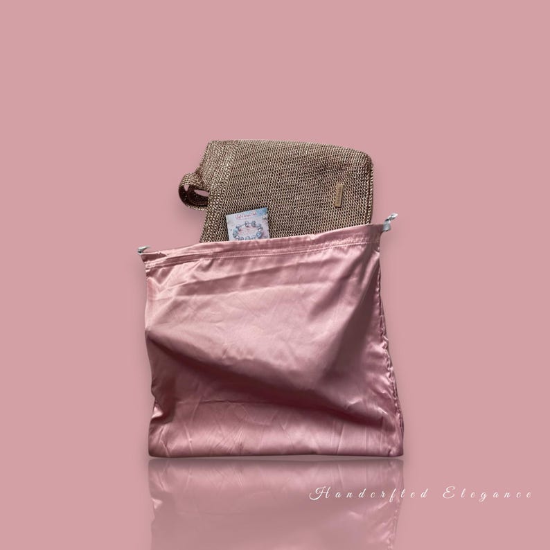 May include: A pink satin drawstring bag with a gold-coloured woven handbag. A small card is visible inside the handbag. The background is a solid pink colour. The words "Handcrafted Elegance" are at the bottom.