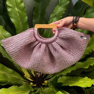 K&ouml;nnte beinhalten: Eine hellrosa geh&auml;kelte Handtasche mit Holzgriff. Die Tasche hat eine abgerundete Form und eine gewebte Textur. Der Griff hat eine hellbraune Farbe. Die Tasche wird vor einem Hintergrund aus gr&uuml;nen Bl&auml;ttern gehalten.