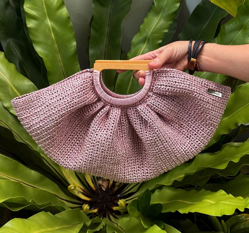 K&ouml;nnte beinhalten: Eine hellrosa geh&auml;kelte Handtasche mit Holzgriff. Die Tasche hat eine abgerundete Form und eine gewebte Textur. Der Griff hat eine hellbraune Farbe. Die Tasche wird vor einem Hintergrund aus gr&uuml;nen Bl&auml;ttern gehalten.