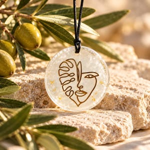 May include: A circular resin pendant necklace featuring a gold-toned line art illustration of a human face merged with a monstera leaf. The pendant has a translucent, pearlescent white base with embedded gold flakes and hangs from a black cord.