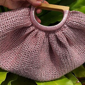 K&ouml;nnte beinhalten: Eine handgefertigte, altrosa geh&auml;kelte Handtasche mit Holzgriff. Die Tasche hat eine runde Form mit einem plissierten Design und einem kreisf&ouml;rmigen Griff. Der Griff ist aus hellem Holz gefertigt. Die Tasche steht vor einem Hintergrund aus gr&uuml;nen Bl&auml;ttern.