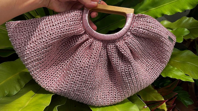 K&ouml;nnte beinhalten: Eine handgefertigte, altrosa geh&auml;kelte Handtasche mit Holzgriff. Die Tasche hat eine runde Form mit einem plissierten Design und einem kreisf&ouml;rmigen Griff. Der Griff ist aus hellem Holz gefertigt. Die Tasche steht vor einem Hintergrund aus gr&uuml;nen Bl&auml;ttern.