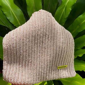 May include: A beige knitted beanie hat with a ribbed texture. The hat has a small, rectangular tag that reads "handmade" in a green font. The hat is displayed against a backdrop of green leaves.