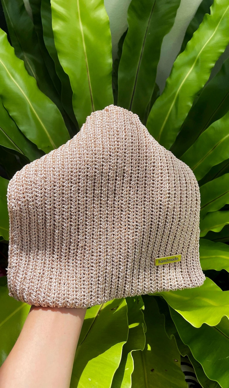 May include: A beige knitted beanie hat with a ribbed texture. The hat has a small, rectangular tag that reads "handmade" in a green font. The hat is displayed against a backdrop of green leaves.