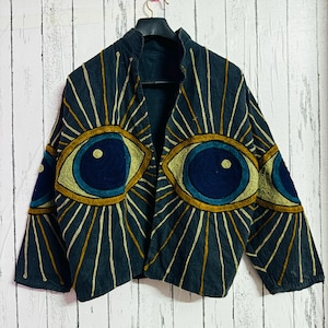 May include: A dark blue jacket featuring embroidered eye designs in gold, yellow, and blue. The jacket has an open front and long sleeves. The eye designs are on the front and sleeves of the jacket.