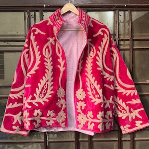 May include: A vibrant fuchsia jacket with intricate gold embroidery. The jacket features a velvet texture and a striped lining. The design includes floral and leaf patterns, creating a decorative and eye-catching garment. The jacket is displayed on a wooden hanger.