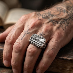Può includere: Un anello d'argento con una faccia rettangolare raffigurante un rilievo religioso dettagliato. L'anello presenta volute ornate sui lati e viene indossato su un dito.