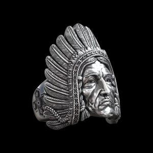 May include: A silver ring featuring a detailed relief of a person wearing a feathered headdress. The design includes intricate textures on the feathers and a patterned band along the front edge, with a geometric motif visible on the side of the ring.