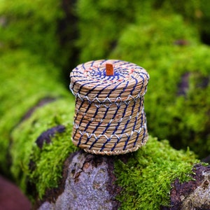 May include: A small, round, woven basket with a blue and white striped pattern. The basket is made of natural materials and has a wooden lid.