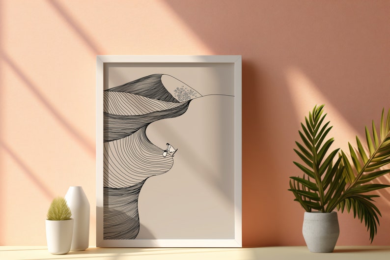 May include: A white framed print with a black and white line drawing of a desert landscape with two figures sitting on a dune. The background is a light beige color.