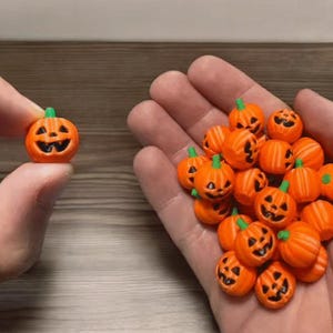 Op de afbeelding: Een verzameling miniatuur oranje pompoenvormige kralen, elk voorzien van een uitgesneden jack-o'-lantern gezicht met zwart geschilderde ogen, neus en mond, en een klein groen steeltje aan de bovenkant, gepresenteerd in een groep en als enkel stuk.
