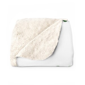 Puede incluir: Una manta doblada que presenta una superficie superior blanca lisa con un pequeño acento circular verde y un forro de forro polar sherpa de color crema grueso y texturizado en la parte inferior. La manta está doblada para mostrar ambos lados.
