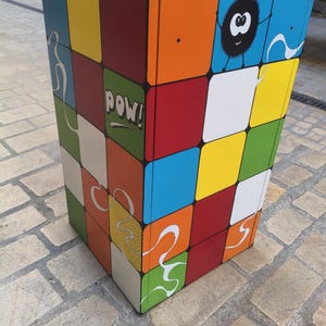Könnte beinhalten: Ein farbenfroher, rechteckiger Mülleimer mit einem Patchwork-Design aus Quadraten in verschiedenen Farben, darunter Rot, Blau, Orange und Grün. Das Wort "pow!" ist auf einer Seite aufgedruckt, und eine Cartoonfigur ist auf einer anderen.