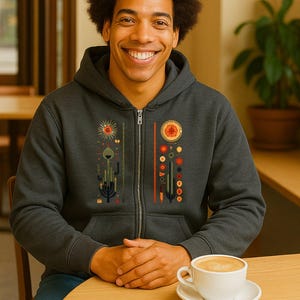 May include: Charcoal grey zip-up hoodie with a desert-inspired design. The graphic includes stylised cacti, suns, and geometric shapes in red, orange, and green. A person is wearing the hoodie while seated at a table with a coffee cup.